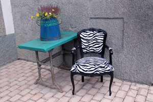 Skickligt hantverk i form av en svartlackerad stol av äldre modell klädd i zebramönstrat tyg vid en bänk med en blomkruka med texten Bagarmossens tapetserare
